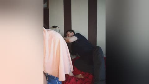A video of a man in a dark shirt, lying on a bed with red sheets, surrounded by vertical brown and white striped walls.