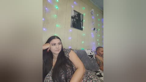 Video of a young woman with long black hair, wearing a floral dress, sitting on a couch with a gray blanket and a man in the background, in a dimly lit room with string lights and a framed photo on the wall.