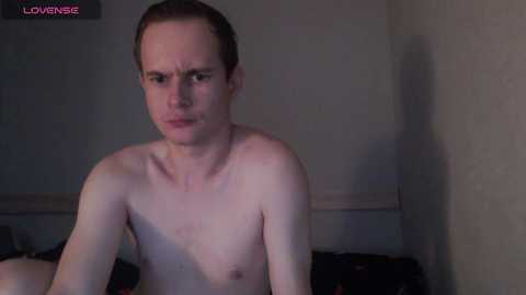 Video of a skinny, fair-skinned, red-haired man with a stern expression, shirtless, sitting in a dimly lit room with beige walls, possibly a bedroom.