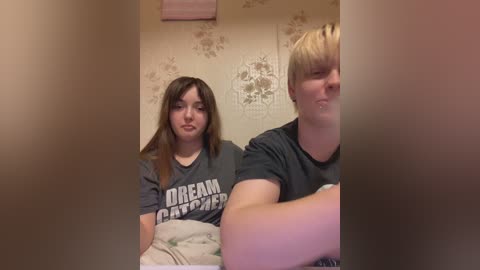 Media: Video of two young women, one with long brown hair, the other with short blonde hair, both wearing grey T-shirts with \"DREAM CATCHER\" text. They are indoors with floral wallpaper and a pink towel in the background.