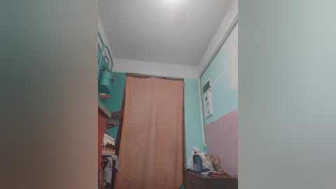 Video of a small, cramped bedroom with light teal and pastel pink walls. A large orange curtain partially covers a window. A dark wooden dresser is cluttered with items, including a blue bottle.