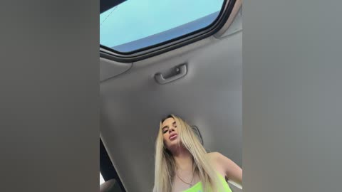 Media: Video of a blonde woman in a neon green top sitting in a car, looking down. The car's interior is modern with a grey dashboard and a sunroof.