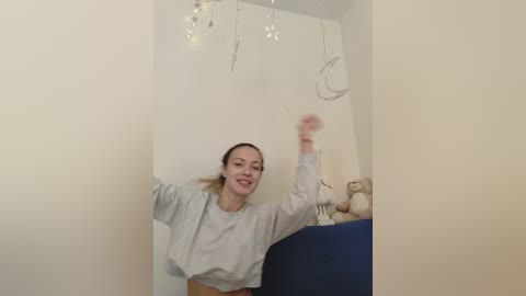 Media: A video of a joyful woman with light skin, brown hair in a ponytail, wearing a light grey sweater, standing in a dimly lit room with white walls, a blue sofa, and a teddy bear.