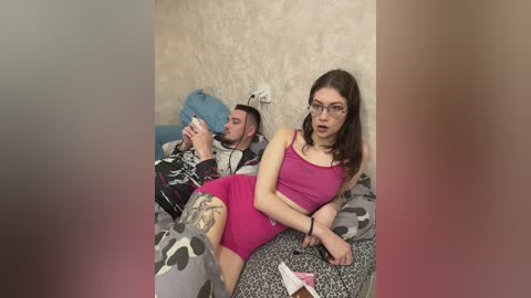 Media: Video of a young woman in pink athletic wear, sitting on a bed with a man, both holding phones, in a dimly lit room.