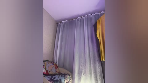 A video of a small, dimly lit room with a light gray curtain, a colorful patterned blanket, and a yellow garment hanging on the wall.