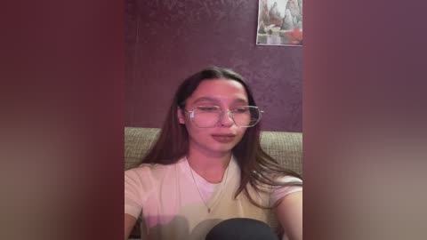 A video of a young woman with long, straight, dark hair, wearing glasses and a beige sweater, sitting on a green couch against a purple wall with a framed picture in the background.