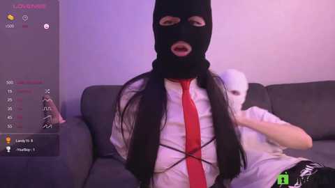 Video of a person wearing a black ski mask and a red tie over a white shirt, sitting on a gray couch, with a white mask on the right and a digital overlay showing a game interface.