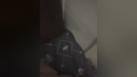 A dimly lit video shows a person wearing a dark, patterned shirt with white symbols. The background is shadowy, with blurred, indistinct objects. The image is grainy, suggesting low-quality or poor lighting.