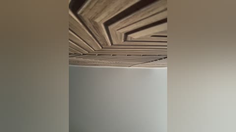 A close-up video of a modern ceiling with geometric, light wood paneling, creating a textured, zigzag pattern. The background is a smooth, off-white wall, emphasizing the intricate design of the ceiling.