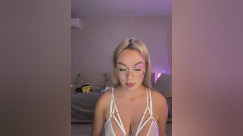 Media: A video of a blonde woman with glasses and a white strappy lingerie top, standing in a dimly lit bedroom with a bed and nightstand in the background.