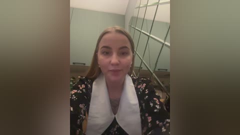 Media: Video of a young woman with fair skin, blonde hair, and a white floral blouse, standing indoors near a glass wall.