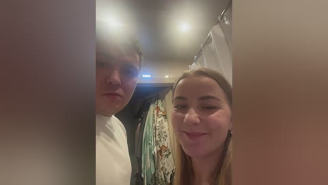 Media: Video of a smiling blonde woman with a white shirt, standing in a dimly lit bathroom with a white shower curtain and a colorful towel hanging.
