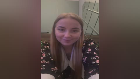 Media: A video of a young woman with straight, light brown hair, smiling. She's wearing a black floral dress and is indoors with a green wall and mirror in the background.