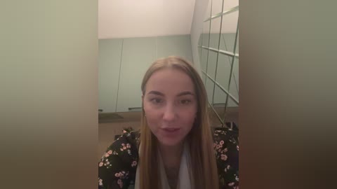 Media: Video of a young woman with long blonde hair, fair skin, and a light floral dress, standing in a modern, minimalist hallway with mirrored walls.