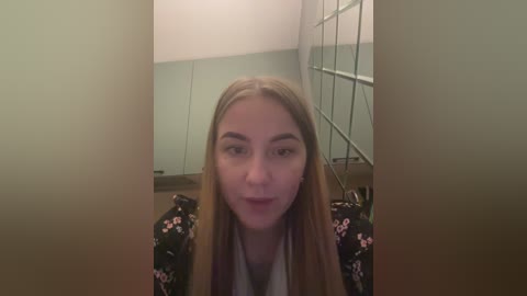 Media: A video of a young Caucasian woman with long, straight blonde hair, wearing a black floral-patterned sweater, standing in a modern, minimalist room with glass walls and white walls.