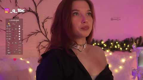 Video of a young woman with light skin, long brown hair, and green eyes, wearing a black off-shoulder top, standing in a cozy room with warm lighting, string lights, and a tree.