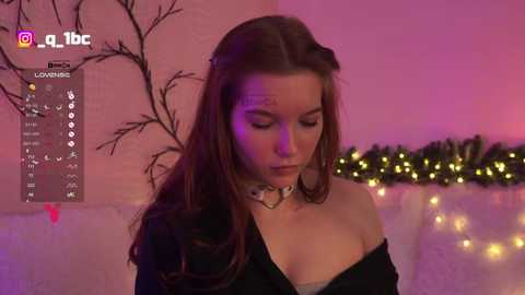 Video of a young woman with long brown hair, wearing a black off-shoulder top, standing against a pink background with glowing fairy lights, and a tree silhouette.