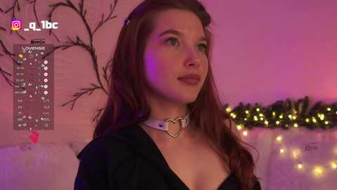 A video of a young Caucasian woman with long, wavy brown hair, wearing a black jacket and a heart-shaped choker, against a pink and purple-lit backdrop with fairy lights.