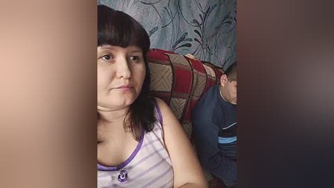 A video of a woman with straight dark hair and a purple tank top sitting on a couch with a patterned cushion. A man in a blue shirt is partially visible behind her.