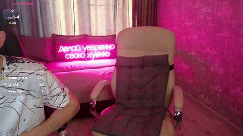 Video of a man in a white T-shirt sitting on a beige recliner with a gray pillow, illuminated by pink neon lights in a dimly lit room with a brick wall and a couch.
