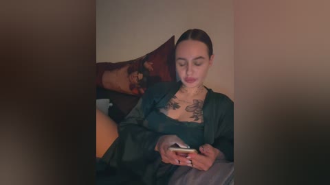 Media: Video of a fair-skinned woman with dark hair, wearing a green satin robe, holding a smartphone, sitting on a bed with dark pillows and a beige wall in the background.