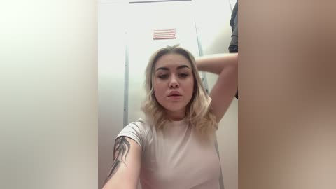 Media: Video of a blonde woman with medium-sized breasts, wearing a white t-shirt, in a public restroom, leaning against a stall door, with a visible tattoo on her arm.