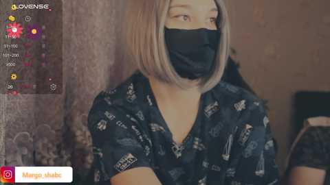 Video of a young woman with light skin, straight shoulder-length blonde hair, wearing a black face mask, navy floral-patterned blouse, and a black lanyard. Background shows a dimly lit room with a textured wall.