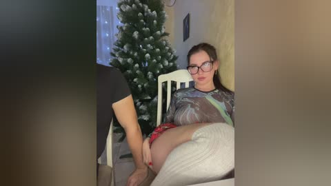 Media: Video of a woman in glasses and patterned shirt sitting on a chair next to a decorated Christmas tree, with a man's legs visible in the foreground.