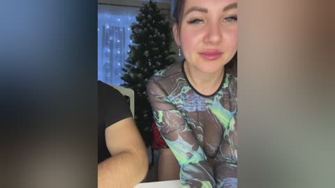 Media: A video of a smiling, light-skinned woman with blue eyes, wearing a sheer, green and black patterned top, leaning forward on a table. Background features a Christmas tree and string lights.