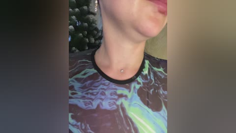 Media: Video of a woman's partially cropped face and torso wearing a vibrant, abstract-patterned shirt with a silver belly button ring. Background includes a green Christmas tree and a blurry, beige wall.