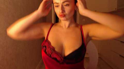 Media: Video of a light-skinned woman with long brown hair, wearing a red lace-trimmed bralette, adjusting her hair in a dimly lit room.