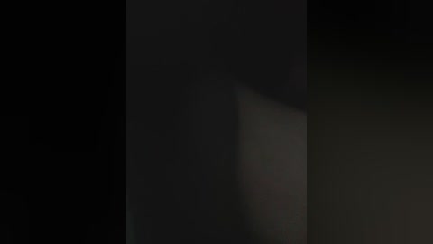 A dimly lit video captures a dark, shadowy silhouette of a person standing against a black background. The figure is indistinct, with only vague outlines and minimal details discernible.