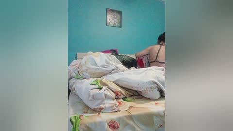 Video of a woman with black hair wearing a bra, lying on a bed with floral sheets in a turquoise room, blurred background.