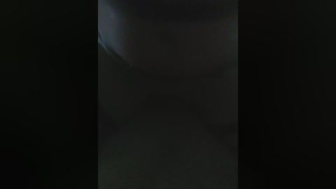 A dimly lit video showing the silhouette of a person with a dark, possibly hooded, top, standing against a dark background, with indistinct features and unclear details due to the low light.