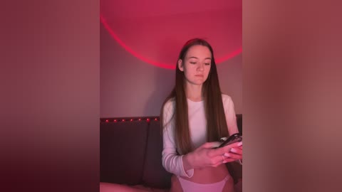 Media: Video of a young woman with long brown hair, wearing a white crop top and white panties, sitting on a black leather couch, holding a smartphone, in a dimly lit room with red ambient lighting.