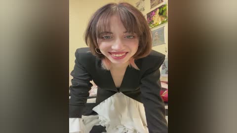 Media: Video of a young woman with shoulder-length brown hair, smiling, wearing a black and white maid outfit, standing in a colorful, cluttered room with floral decorations.