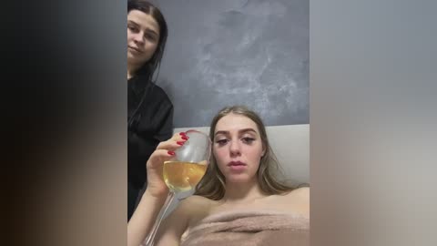 Media: Video of a woman in a spa, with another person behind her, holding a wine glass. The woman is reclining on a massage table, wrapped in a towel, with a serene expression.