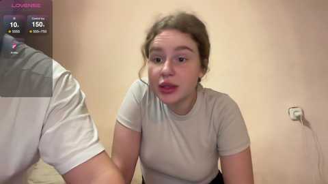 Media: A candid video of a young woman with light skin, brown hair, and a surprised expression, wearing a light grey t-shirt, sitting on a bed in a beige room.