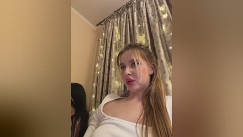 Video of a young Caucasian woman with fair skin and long blonde hair, wearing a white top, standing in front of a window with sheer curtains and fairy lights.