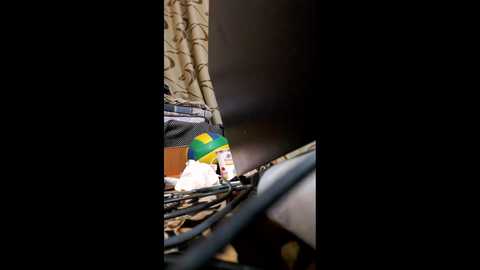 Video of a cluttered desk with a beige, patterned curtain in the background. The desk is messy, featuring a laptop, scattered papers, a coffee cup, and a green and yellow pen cap. The scene is dimly lit, emphasizing the chaos.
