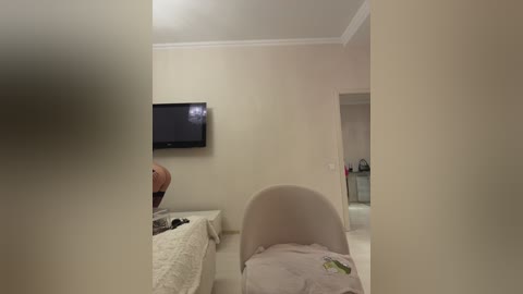 Video of a dimly lit, minimalist bedroom with beige walls, a flat-screen TV mounted on the wall, a white chair, and a bed with white bedding.