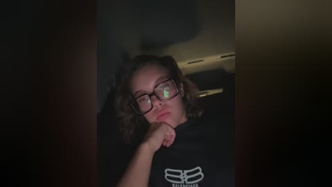 Media: Video of a young woman with curly brown hair, wearing black-framed glasses, and a black t-shirt with a white logo. She rests her chin on her hand, looking contemplative in a dimly lit room.