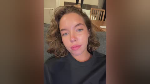 Media: Video of a young woman with shoulder-length wavy brown hair, fair skin, and light makeup, wearing a black t-shirt, sitting indoors on a grey couch with wooden furniture and chairs in the background.