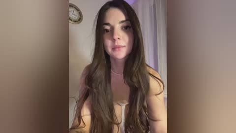 Media: Video of a young woman with long brown hair, light skin, and a slender build, wearing a white top. She stands in a beige room with a clock and a white curtain in the background.