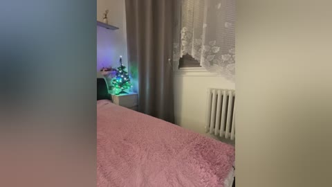 Video of a cozy bedroom with a pink bedspread, white lace curtains, a small Christmas tree, and a radiator under a window, bathed in soft, ambient lighting.