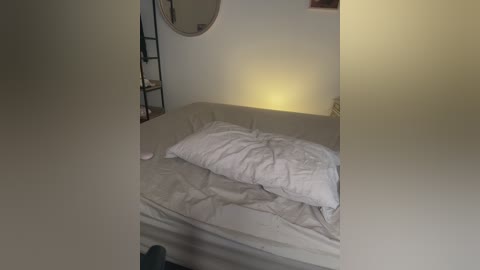 Video of a dimly lit, cluttered bedroom with a beige sofa and messy white bedding. A round mirror hangs on the wall, and a small shelf holds a few items. The room has a cozy, lived-in feel.
