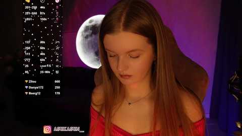Video of a young Caucasian woman with long brown hair, wearing a red off-shoulder top, looking down thoughtfully in a dimly lit room with a moonlit backdrop.