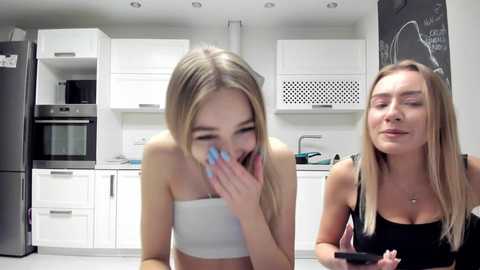 Media: Video of two young women in a modern kitchen: a blonde in a white bandeau top, covering her face with her hands, and a blonde in a black tank top, holding a phone, both with straight hair.