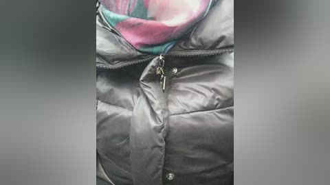 Video of a person wearing a shiny, black puffy jacket with a zippered front and a colorful, patterned scarf peeking out from the collar. The background is a blurred gradient of gray.
