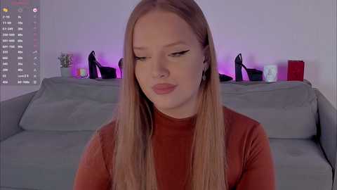 A video of a young, fair-skinned, blonde woman with long, straight hair in a red turtleneck, sitting on a gray couch in a modern living room with purple lighting, high heels, and a TV screen visible.
